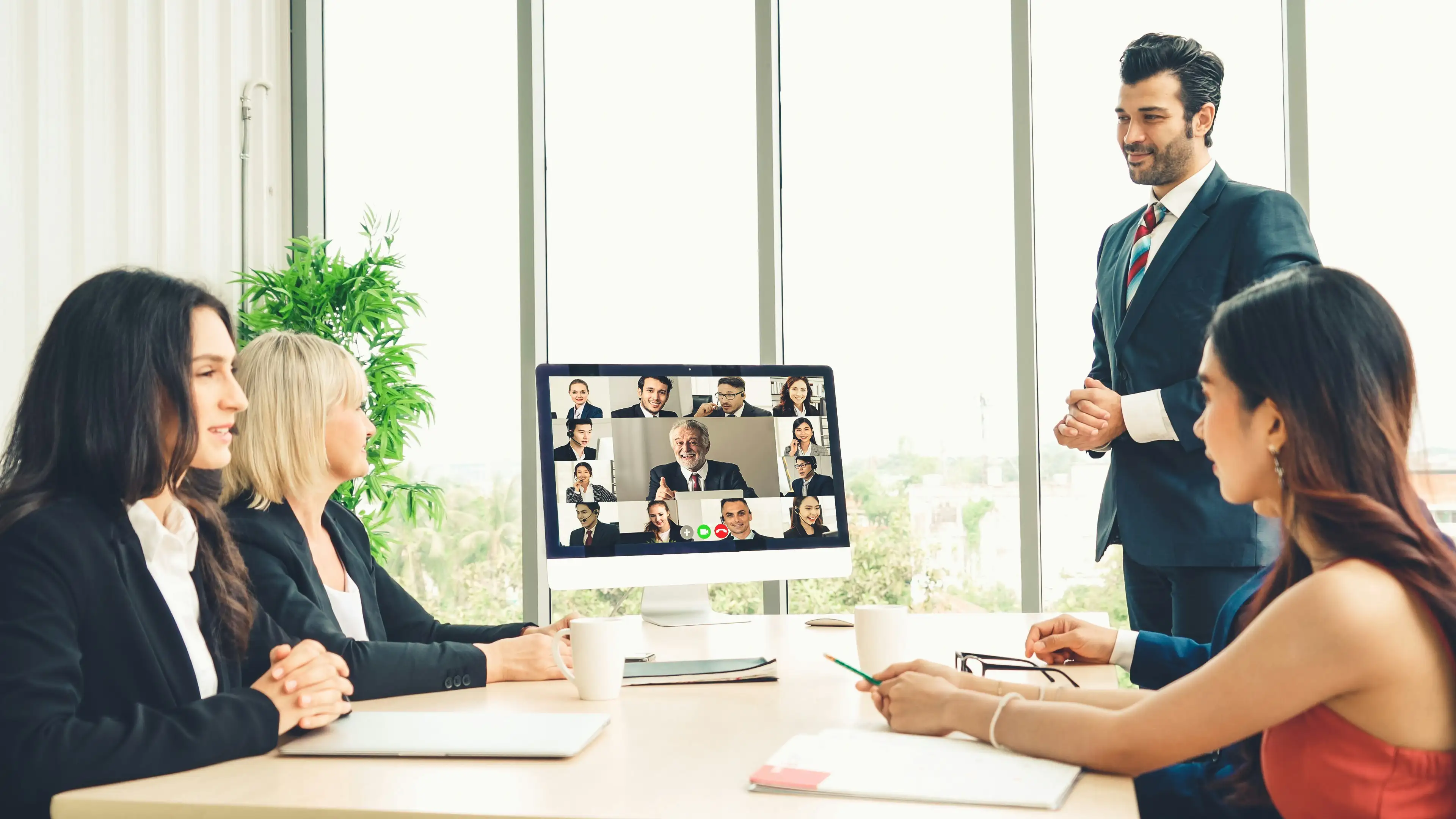Team sitzt gemeinsam am Tisch mit Bildschirm auf dem Online Meeting zu sehen ist
